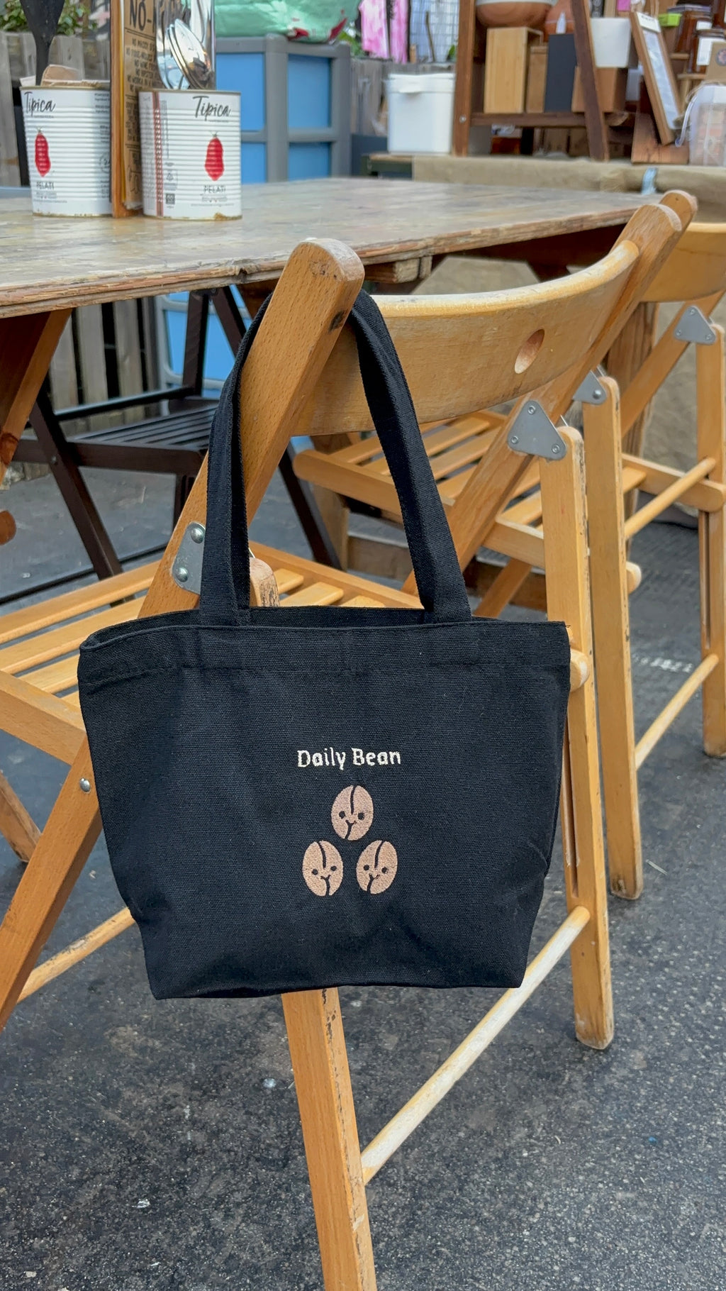 Black tote bag with 'Daily Bean' logo on a wooden chair outdoors.