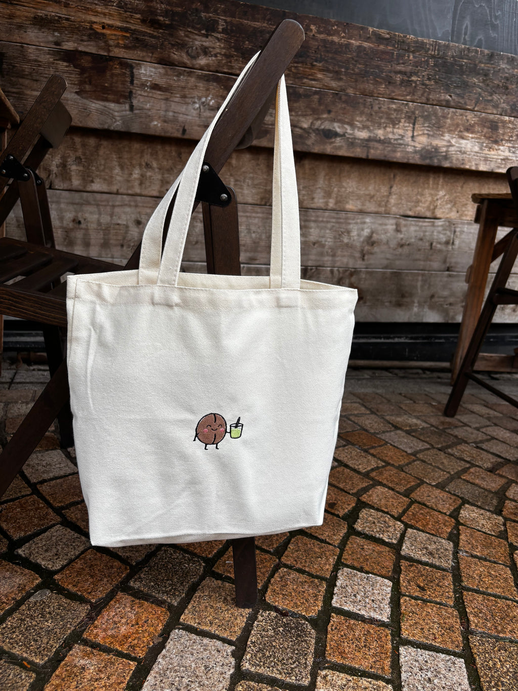 White tote bag with a small design on a wooden chair against a stone wall.