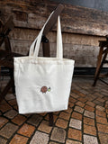 White tote bag with a small design on a wooden chair against a stone wall.