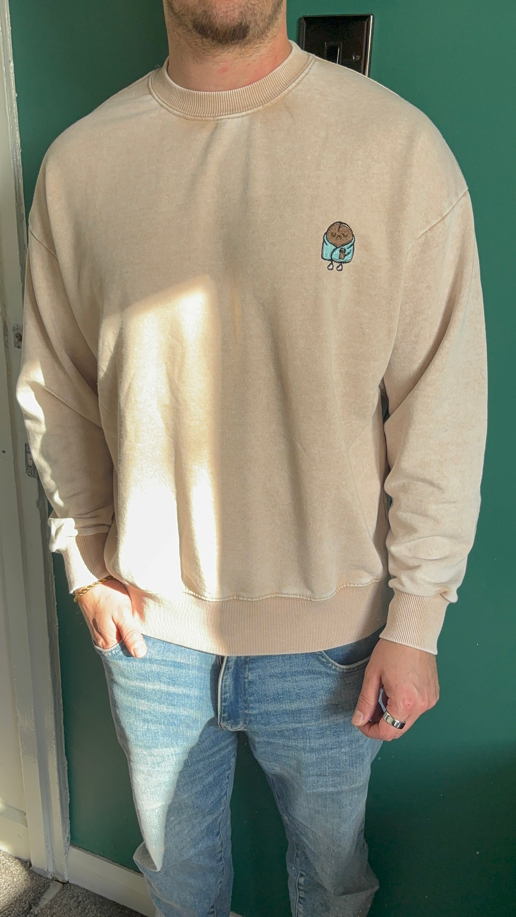 Person wearing a beige sweatshirt with a small graphic on a green wall.
