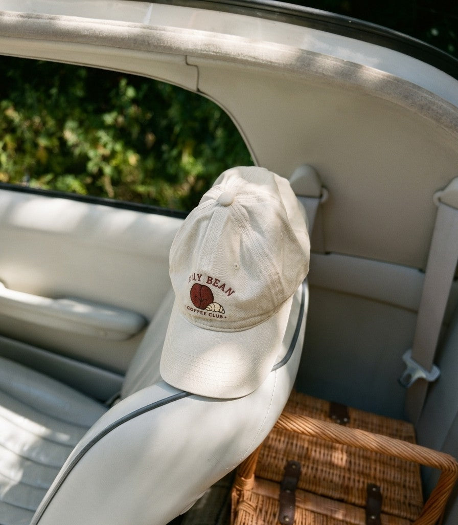 An off-white baseball cap with a brown embroidered logo reading 'DAY BEAN' and 'SANTA MONICA' resting on the back of a car seat inside a vehicle.