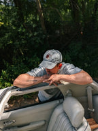 A man wearing a gray baseball cap and a gray t-shirt leans over the open door of a light gray convertible car, with a dense green forest in the background.