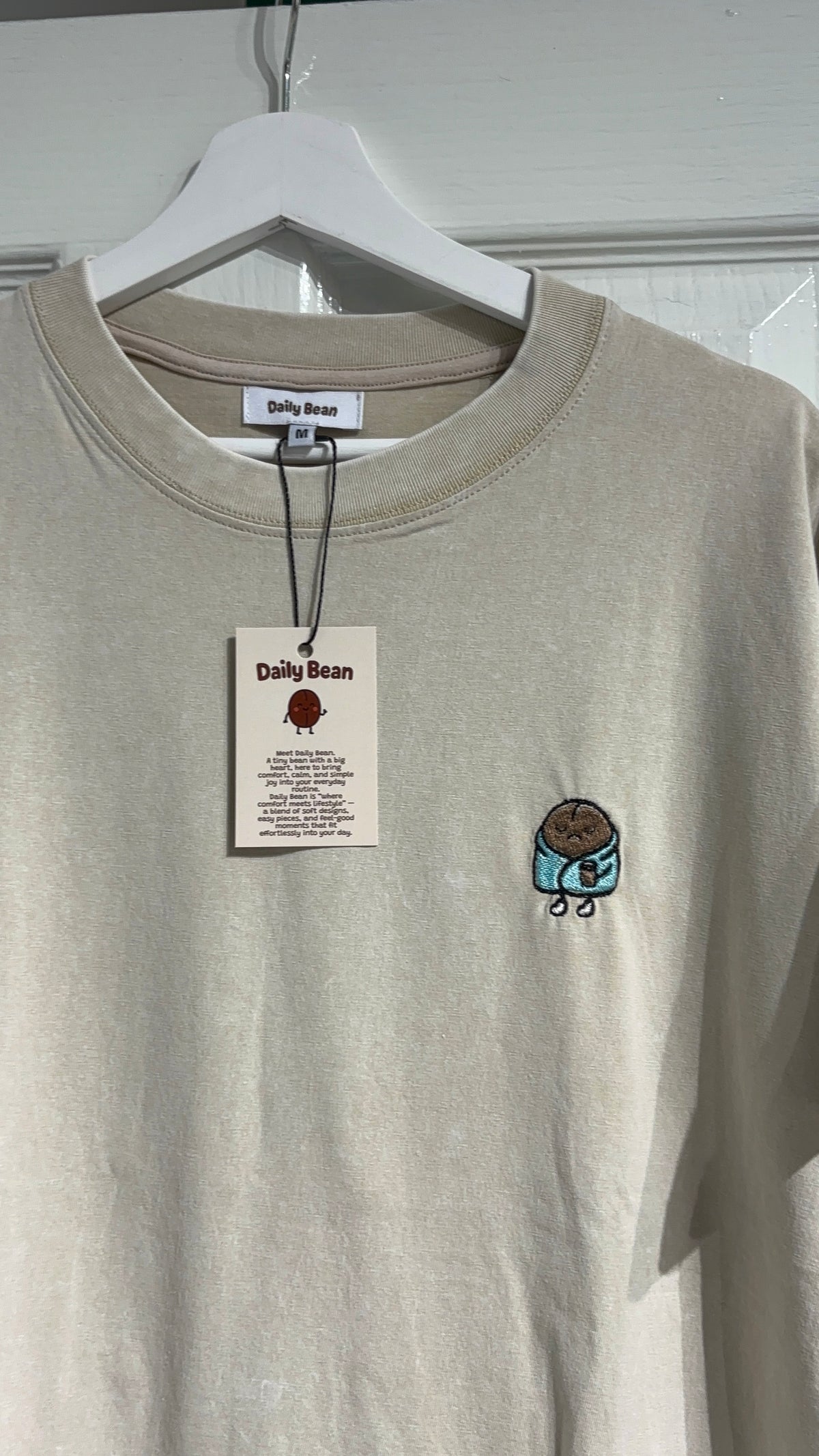 A light beige crewneck sweatshirt from Daily Bean, featuring a small embroidered cupcake graphic on the left chest, hanging on a white hanger with its original paper tag visible.