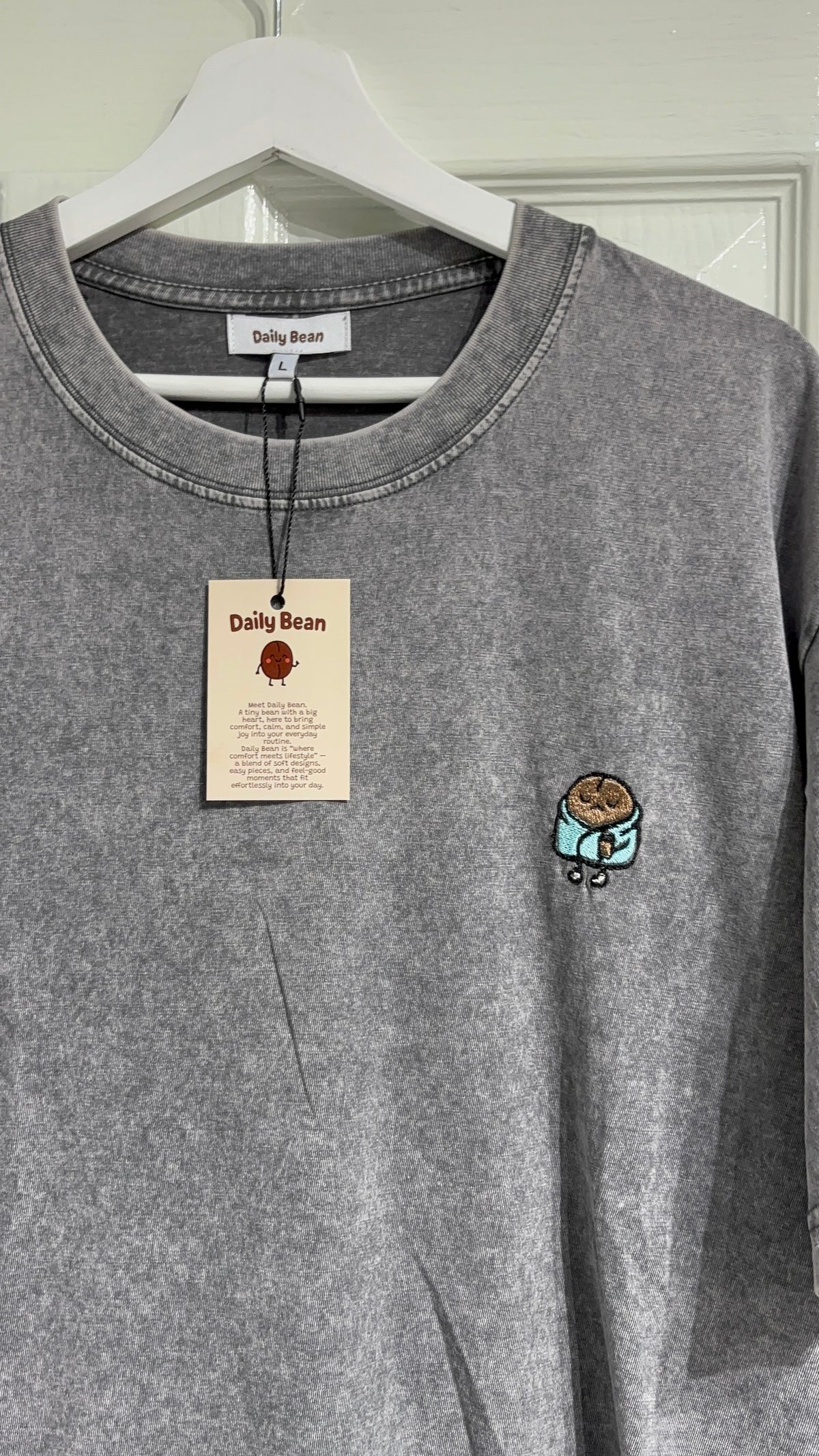 A gray heather crewneck sweatshirt from Daily Bean hanging on a white hanger. The sweatshirt features a small embroidered cartoon character on the left chest and has its original paper tag attached.