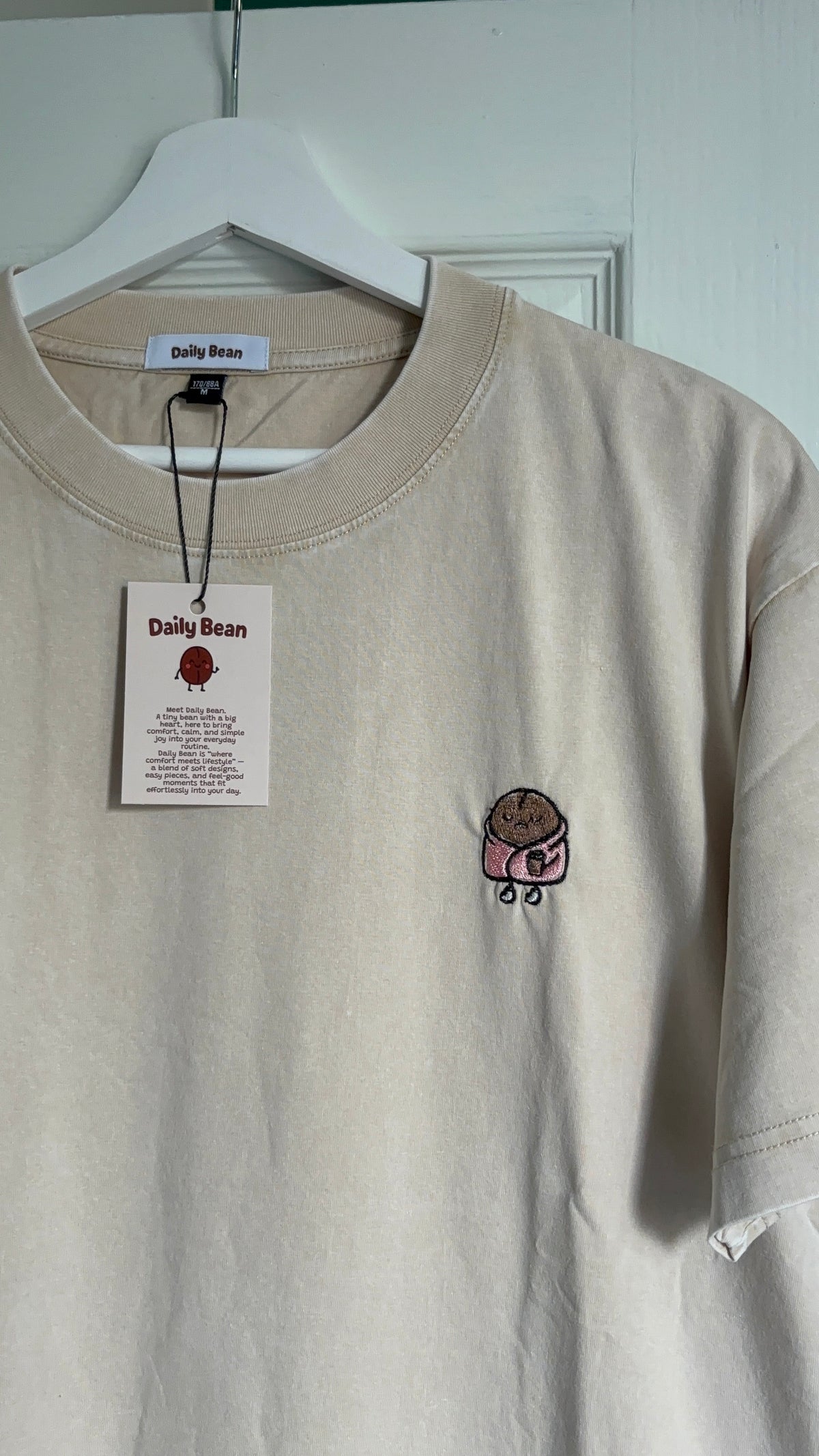 A beige crewneck t-shirt from Daily Bean hanging on a white hanger against a white door. The shirt features a small embroidered cartoon character on the left chest and has its original paper tag attached.