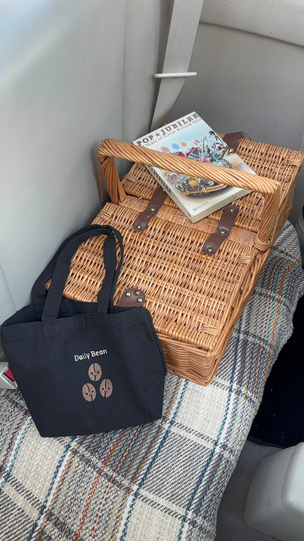 large Black tote bag with a logo next to a wicker picnic basket on a checkered blanket.