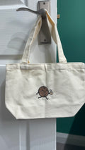 White tote bag with a design on a hook against a white wall.