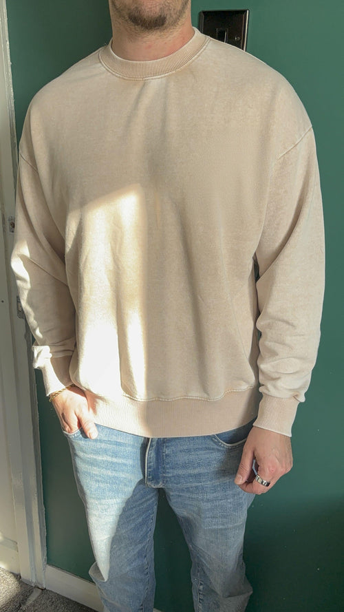 Person wearing a beige sweatshirt and blue jeans against a green wall.