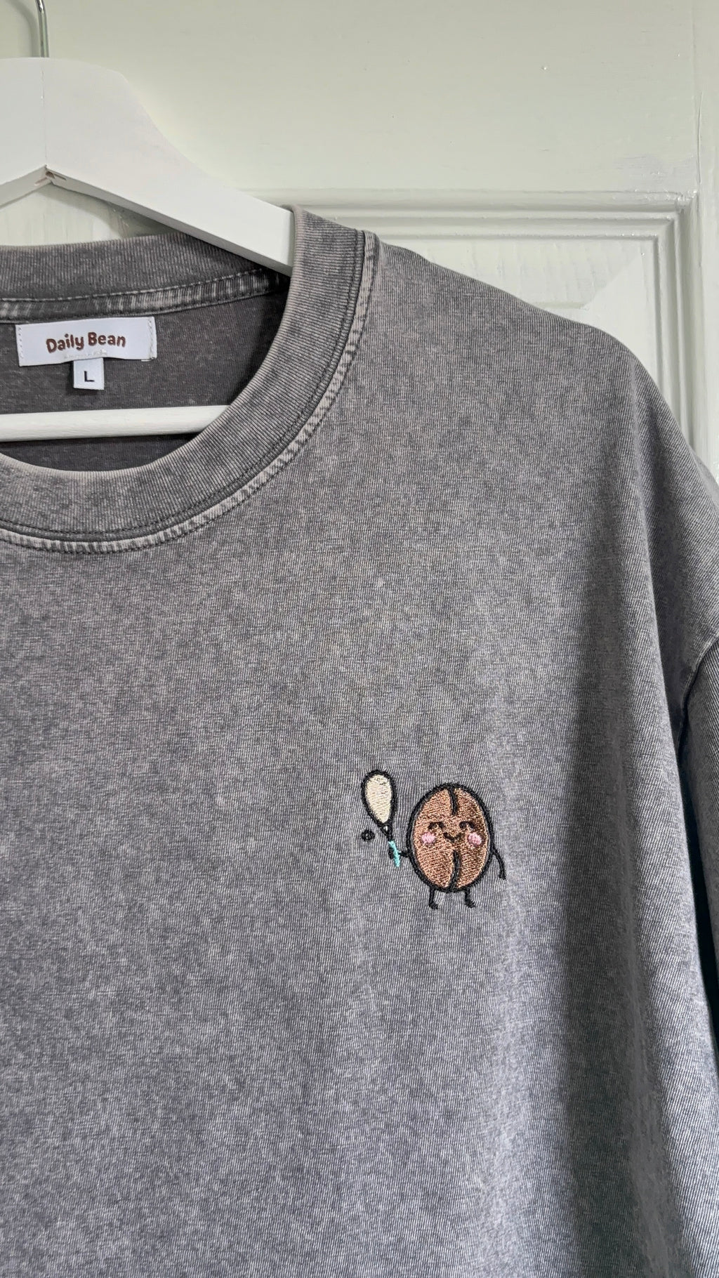 Gray sweatshirt with a small embroidered design on a hanger against a white wall.