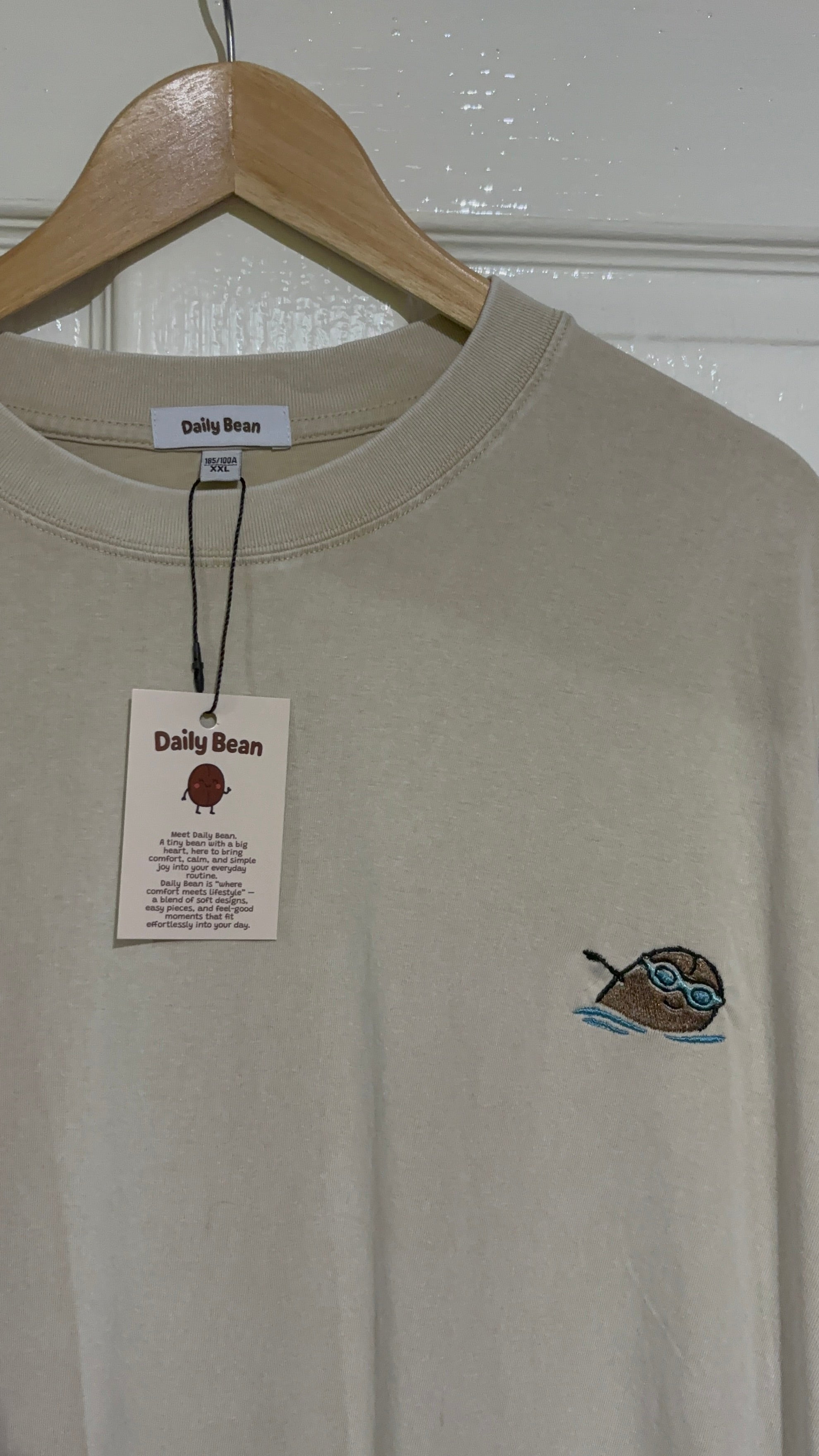 Beige t-shirt with a small logo on a hanger against a white wall.